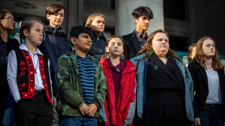 A group of young people stand together outside, on steps in front of a public building.