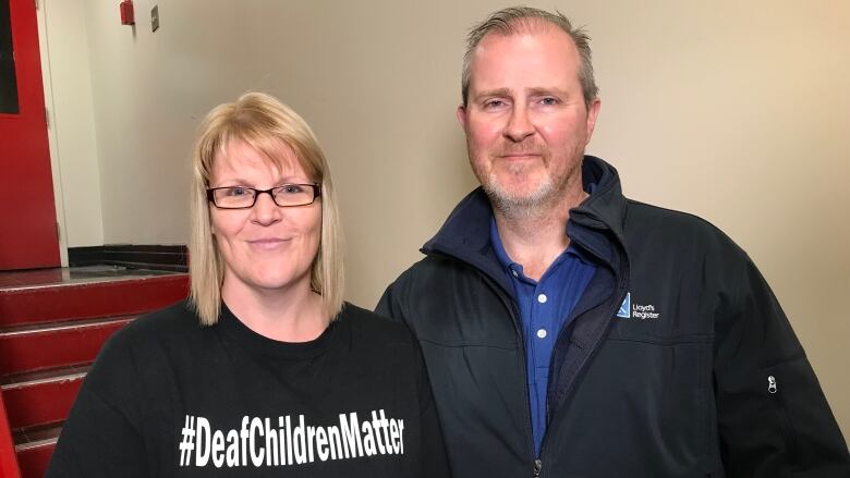 A blonde woman standing next to a grey-haired man. She's wearing a black T-shirt that says Deaf Children Matter. He's wearing a navy blue windbreaker over a royal blue buttom-up shirt.