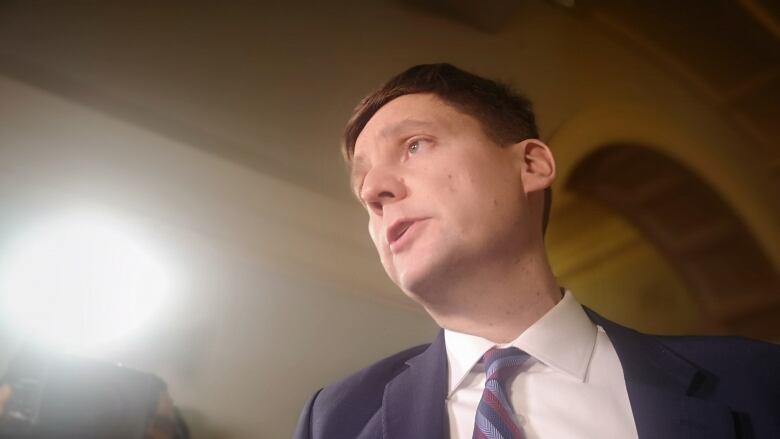 A man in a white shirt, striped tie and a navy suit addresses reporters off-camera under the glare of the television lights.