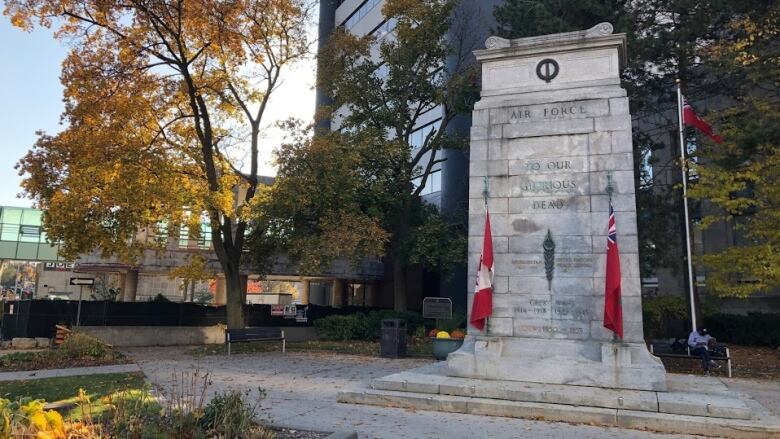 A cement memorial dedicated to soldiers, with a Canadian and Ontario flag in front. The leaves are turning yellow on a tree beside the cenotaph.