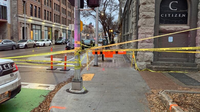 Police tape stretches across a sidewalk and street. Police markers can be seen on the road in the background.