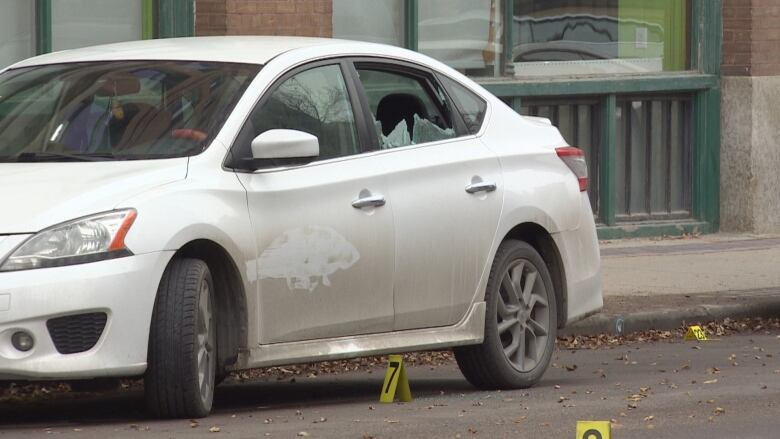 A car with a broken rear passenger window sits on a street with numbered police markers on it.