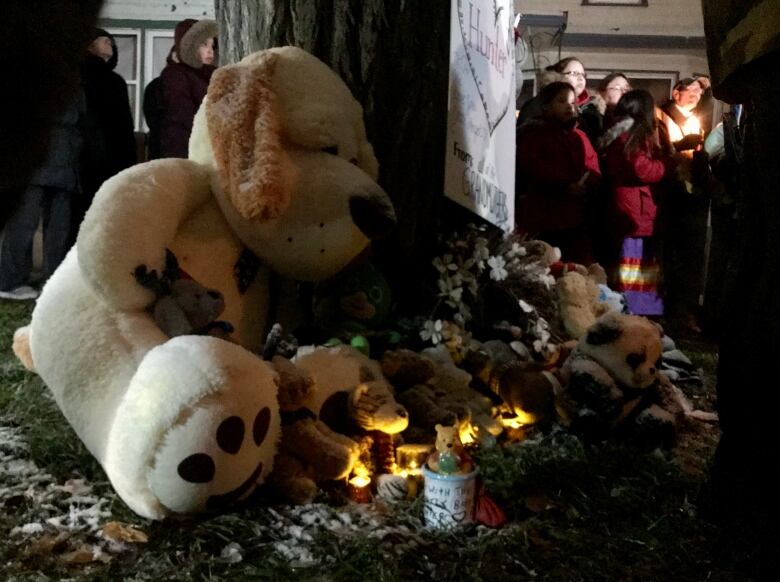 A large stuffed animal in the foreground leans toward a tree surrounded by stuffed animals, with a few small lights among them. People holding lights stand in the background.