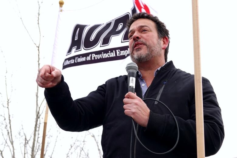 A man with a beard holds a microphone. He's standing in front of a white flag that says AUPE.