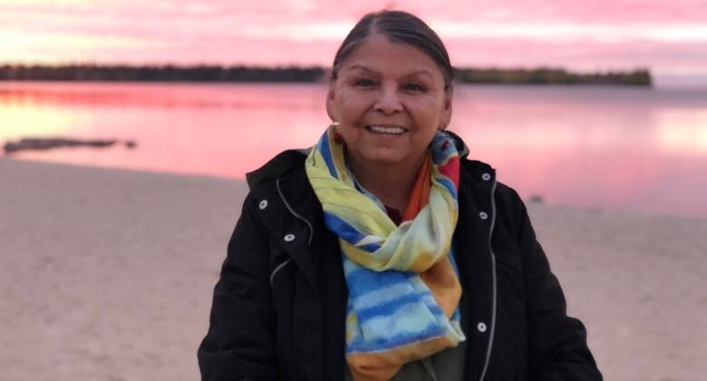 A woman who's smiling is pictured in front of a body of water with a pink sky sunset in the background.