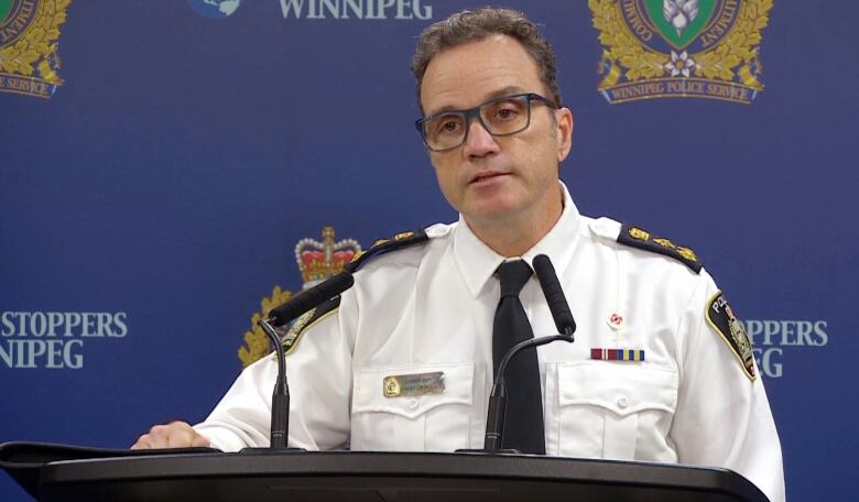 A man in a uniform shirt and tie stands behind a podium with microphones.