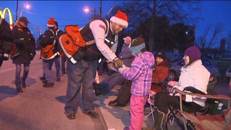 A man wearing a Santa hat and a Canada Post uniform hands candy to a child in a group along the side of a street as a parade passes at twilight.