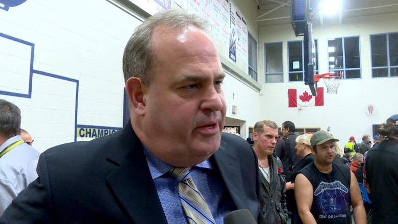 A man in a dark suit and a tie speaks into a microphone in a school gymnasium with other people milling about in the background.