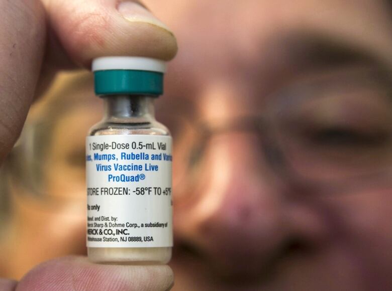 A close-up of a man holding a measles, mumps, rubella and varicella (MMR) vaccine vial between his right thumb and index finger, in front of his face.