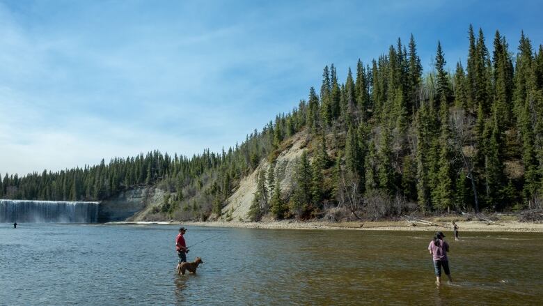 People are seen standing in a river fishing, with a waterfall in the background.