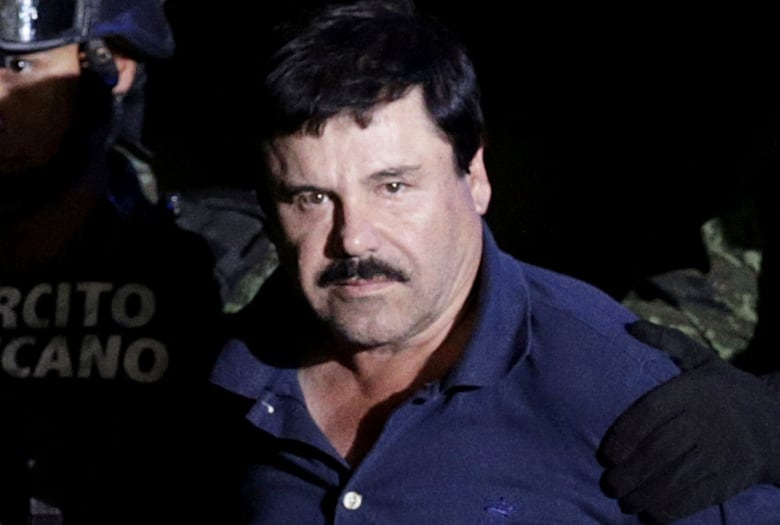 A man with black hair and a moustache, wearing a navy shirt, is escorted by police.