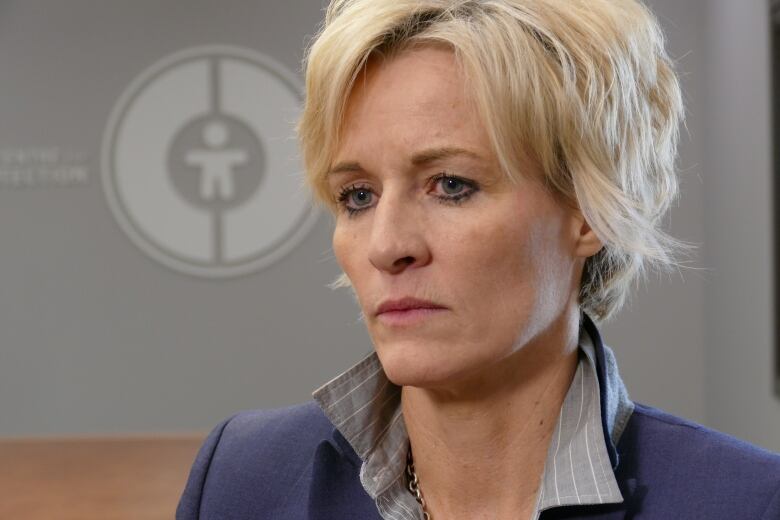 Woman with short blonde hair, grey shirt and blue blazer sits in front of a grey wall.  