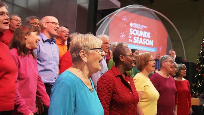 A choir of adults wearing brightly coloured sweaters sing for the audience