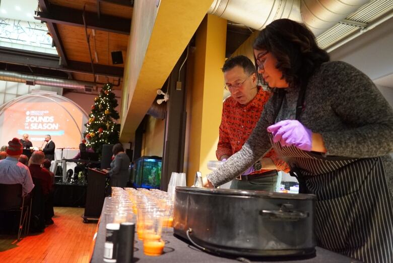 A female chef dishes up a soup from a large pot. Beside her is a man who is interviewing her. A large audience is watching.