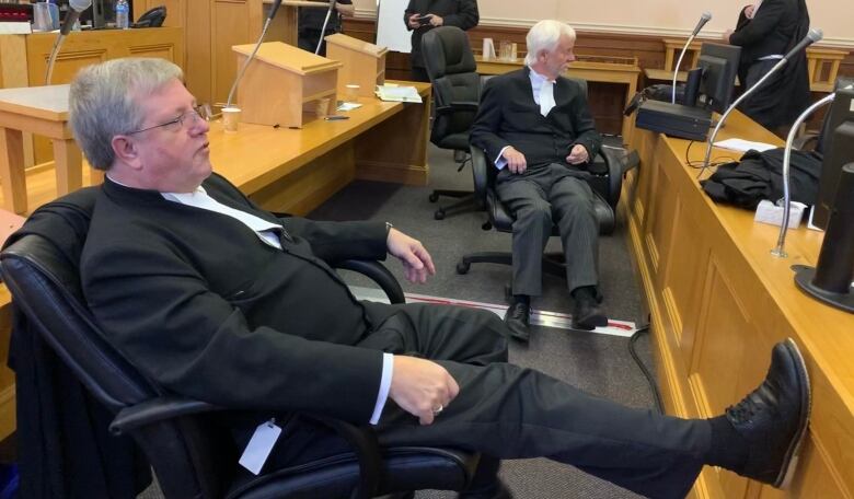 A lawyer in black robes sits with his feet up in a courtroom.