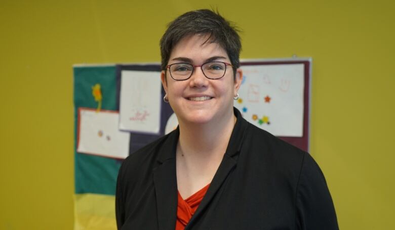 A woman with short, dark hair and glasses poses for a portrait in front of a bulletin board. 