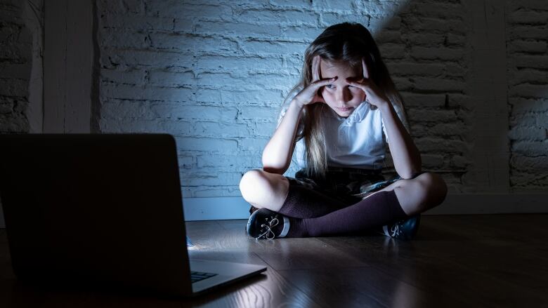 Looking distraught, a young girl sits crosslegged on the floor in a darkened room with her head in her hands, peering at an opened laptop facing her.