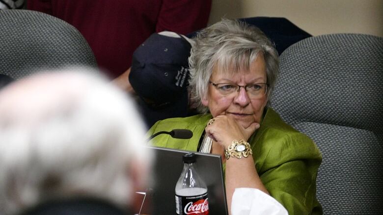 A woman with grey hair weraing a green jacket, glasses and an ornate bracelet sits in a grey chair behind a laptop and a microphone.