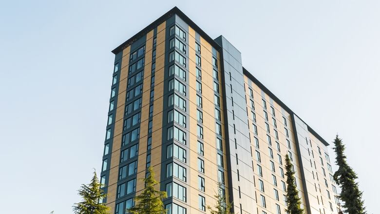 An 18-storey building with a wood exterior is flanked by pine trees. 