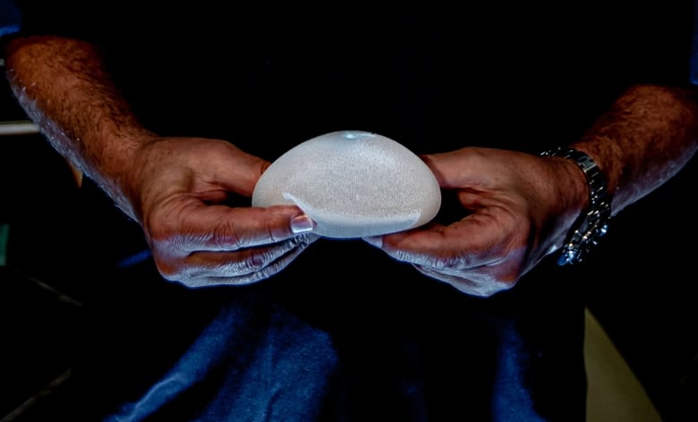 The hands of a plastic surgeon in blue scrubs hold a breast implant.