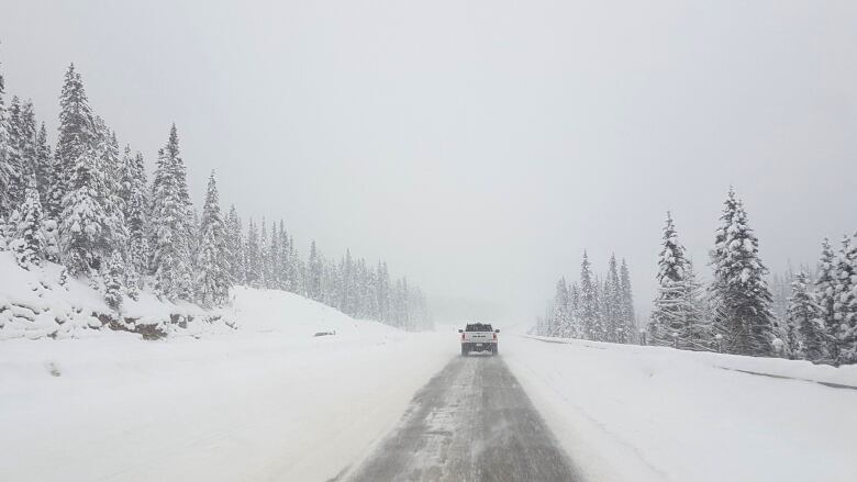 A pickup truck drives along the clear centre lane of a snow-covered highway surrounded by trees as snow falls.