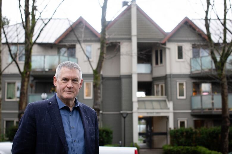 A white man with grey hair poses in front of a 3-storey building.