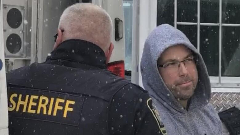 A man wearing glasses and grey roots hoodie stands near a white van on a snowy day.