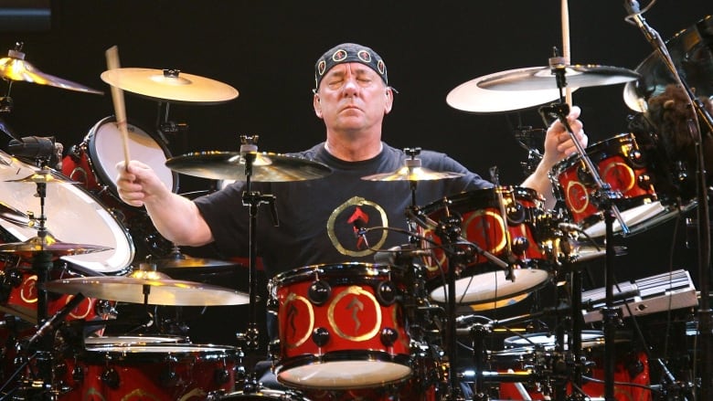 A man sitting at a drum set with his arms stretched wide, while holding drum sticks in each hand, and his eyes closed. 