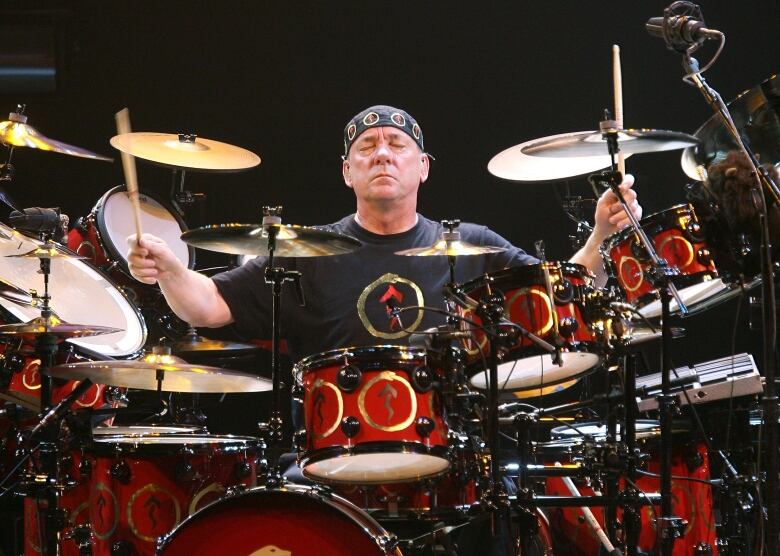 A man sitting at a drum set with his arms stretched wide, while holding drum sticks in each hand, and his eyes closed. 