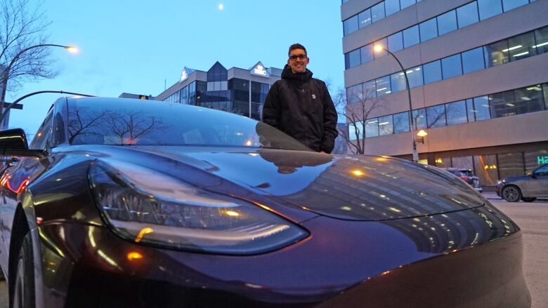 A man stands in downtown Saskatoon beside a Tesla car.