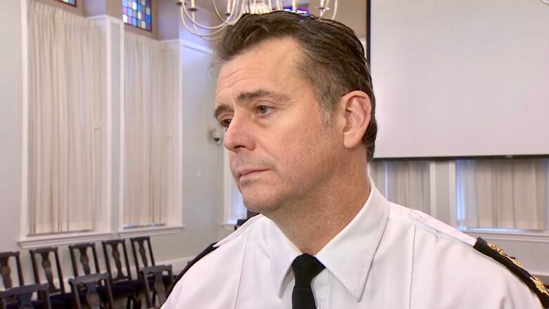 A white man in a police officer's uniform of white, collared shirt and black tie stands in an official-looking ceremonial room at City Hall