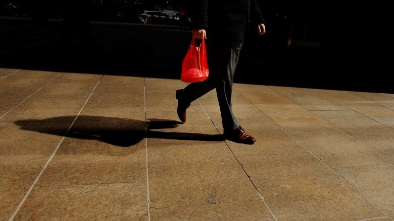 A view of a person's legs as they carry a red plastic bag outdoors.