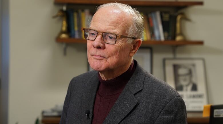 An elderly man is shown looking forward. He is wearing glasses, a blazer and a turtleneck.