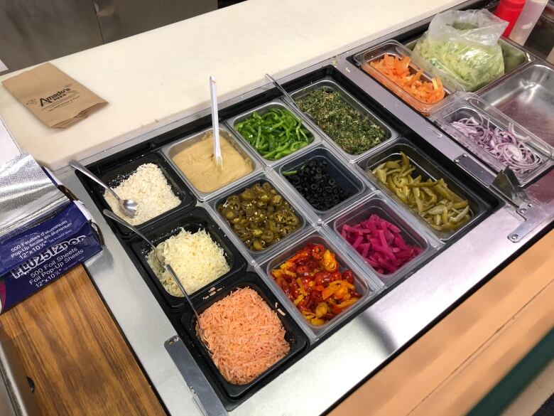 A restaurant counter holds containers of toppings like grated cheese, hot peppers, pickles, olives and sliced onions.