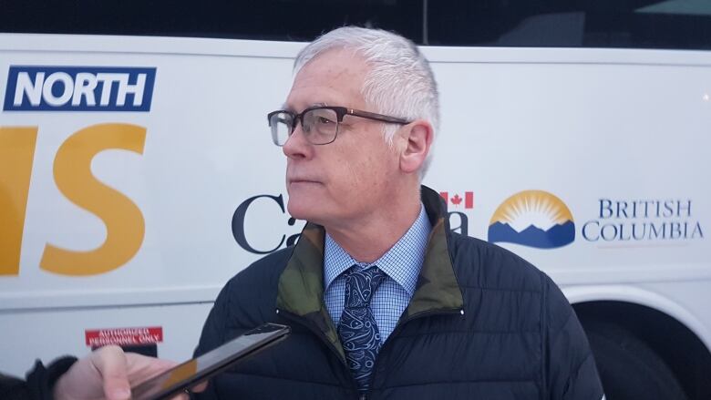 A close-up shot of a man in a dark blue jacket and a striped shirt with a blue tie. A bus is visible in the background.