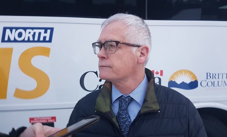 A close-up shot of a man in a dark blue jacket and a striped shirt with a blue tie. A bus is visible in the background.