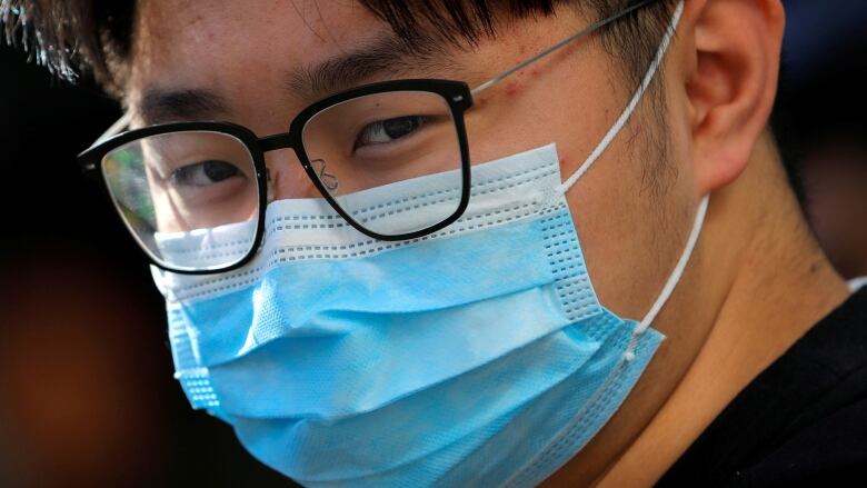 A close-up of a young man wearing glasses and a blue medical mask.