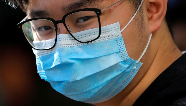 A close-up of a young man wearing glasses and a blue medical mask.