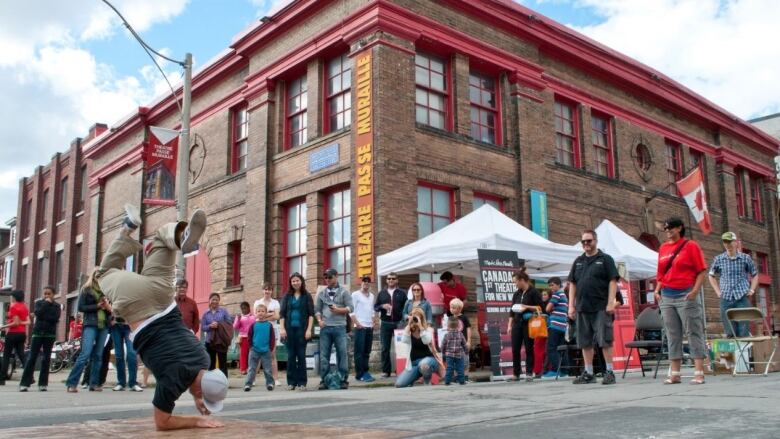 A crowd gathers in front of a brick building to watch a person breakdance.