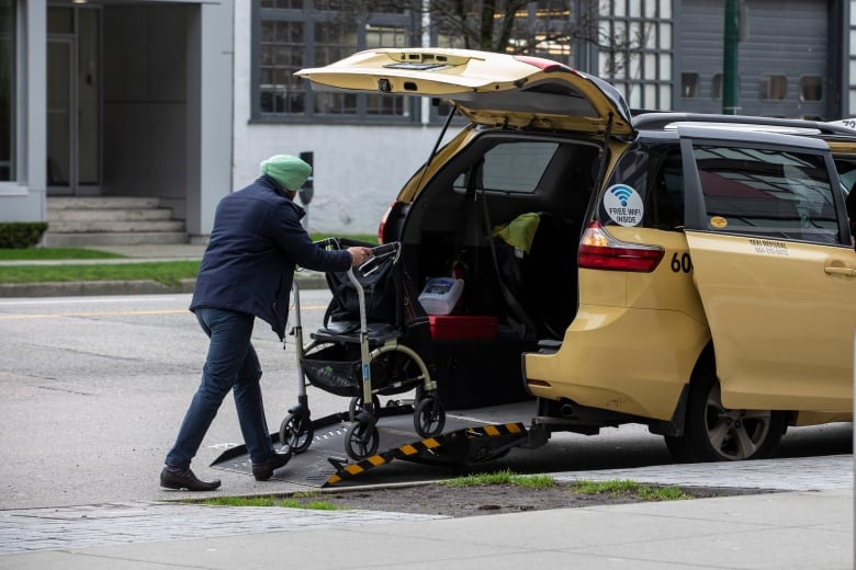 A man wearing a turban wheels a wheelchair into the back of a yellow taxicab.