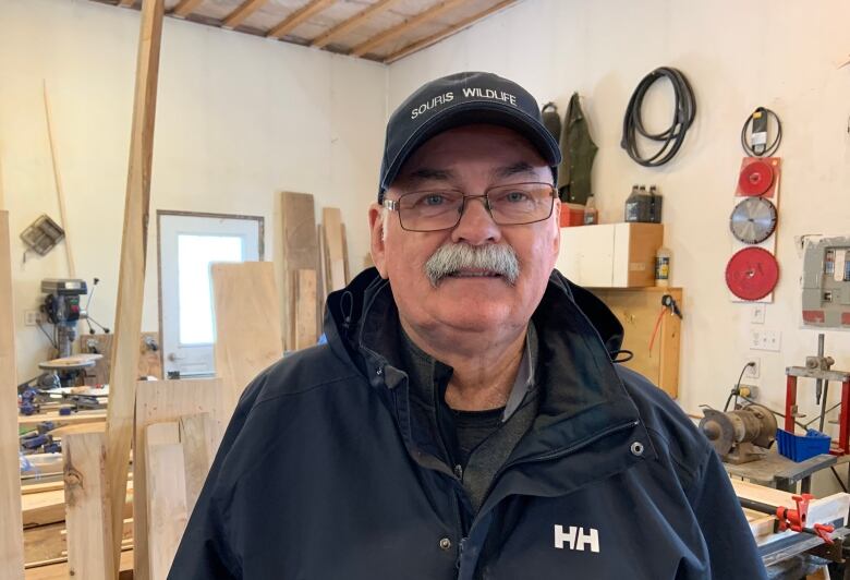 White-haired man with mustache, glasses and ballcap stands in a wood shop. 