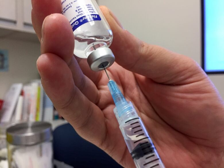 Closeup of a hand holding a bottle of influenza vaccine, with a syringe plunged into it.