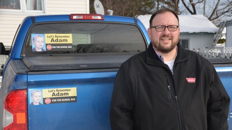 A man stands in front of a blue pickup truck with a Let's Remember Adam bumper sticker.