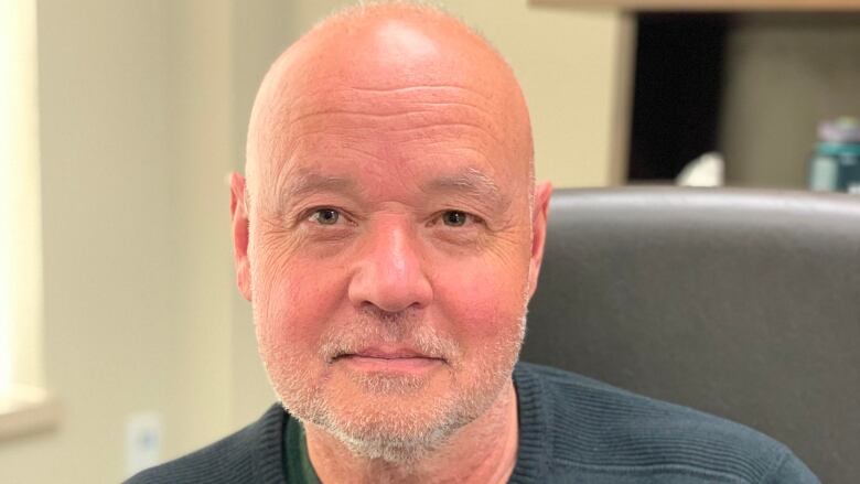 A bald man in a dark, striped sweater sits in an office chair.