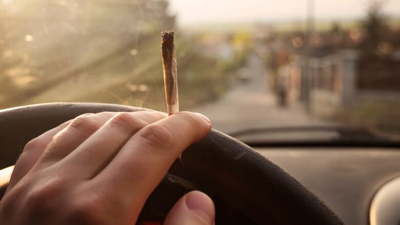 A hand on a steering wheel is also holding a cannabis cigarette between two of the fingers.