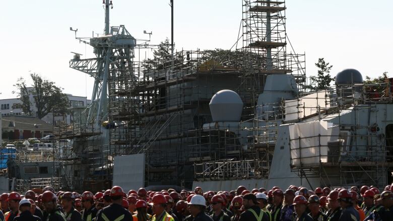 Marine tradespeople gather around and look upon a warship covered in scaffolding.