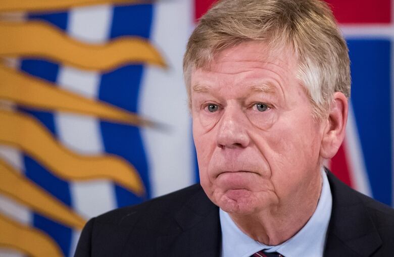 An image of Austin Cullen  a white man with greying hair, dressed in a blue shirt and a navy coat  in front of a flag of British Columbia.