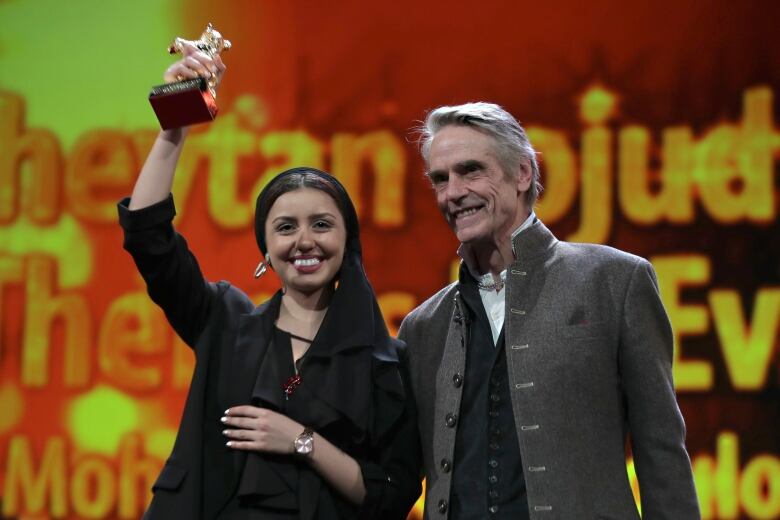 A smiling woman holds up a trophy as she is embraced by a man standing beside her. 