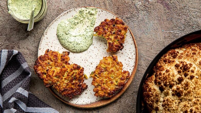 An overhead shot of a white plate with three cauliflower fritters and some green sauce on it. A whole roasted cauliflower is sitting next to it. 