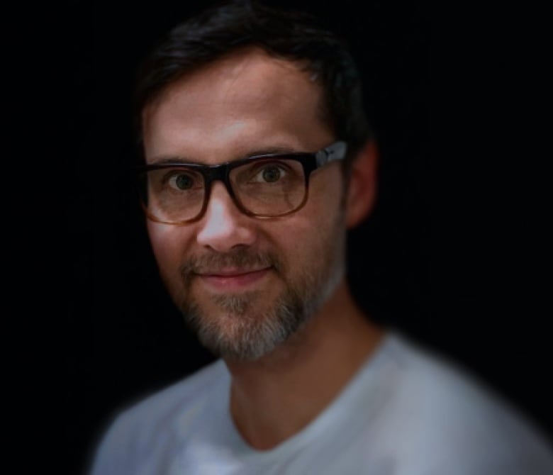 A young man with a goatee wearing a white shirt and glasses smiles at the camera.